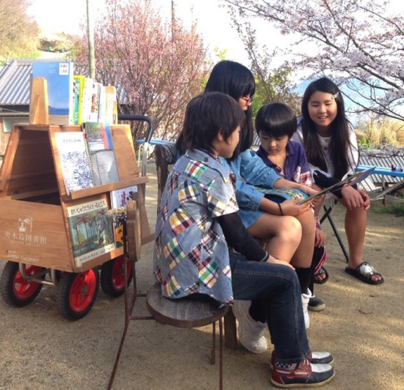 男木島のアーティスト、「オンバファクトリー」制作の手押し車”オンバ”に本を載せて。桜の下、夢中で本を読む子どもたち